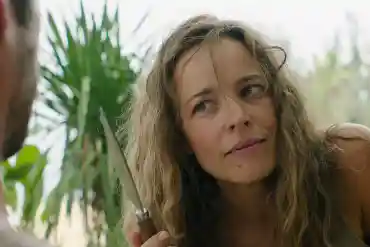 A woman with long, wavy hair looks thoughtfully at a man holding a knife in a lush, green outdoor se.