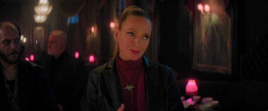 A woman with red hair and a black leather jacket in a dimly lit bar scene.