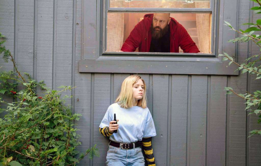 A woman holding a gun outside a house with a man looking through the window.