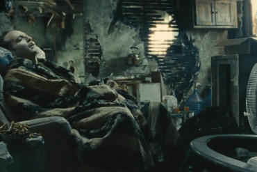 A woman resting in a cluttered, dimly lit room with damaged walls and old furniture.