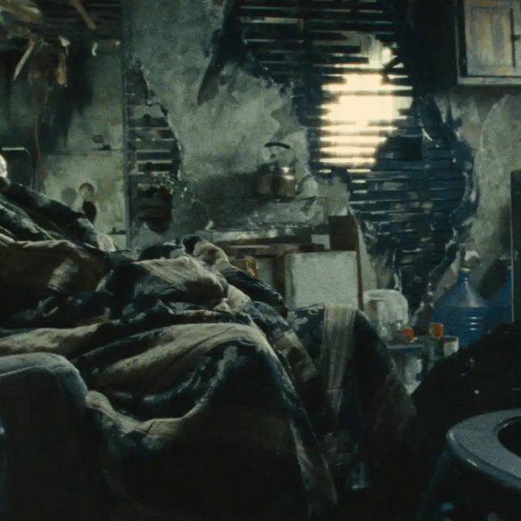 A woman resting in a cluttered, dimly lit room with damaged walls and old furniture.