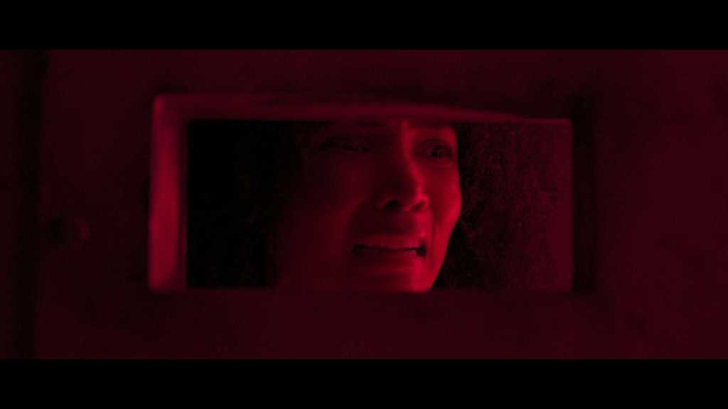 A woman looking distressed behind a mirror with red lighting.