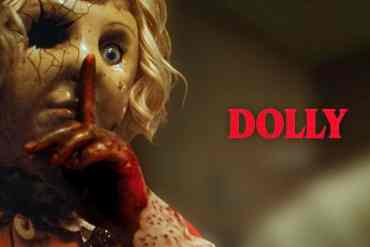 Eerie doll with cracked face and glowing eye, holding finger to lips in a spooky pose.