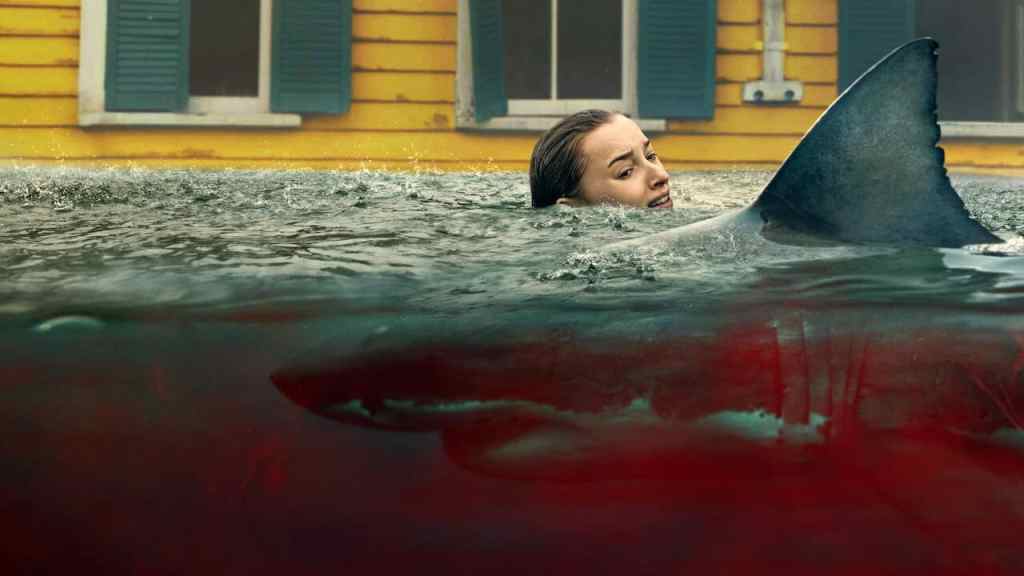 Shark swimming near woman in water with yellow house background.