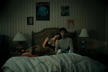 A woman leaning on a man sitting on a bed in a dimly lit bedroom.