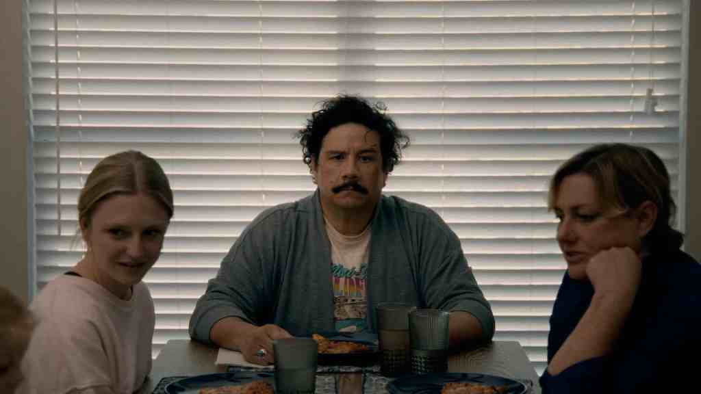 Three people sit at a table with blinds in the background, appearing serious.