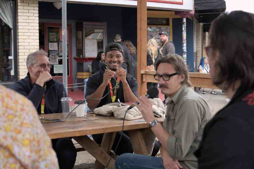 Group of people recording a podcast outdoors at a wooden table.