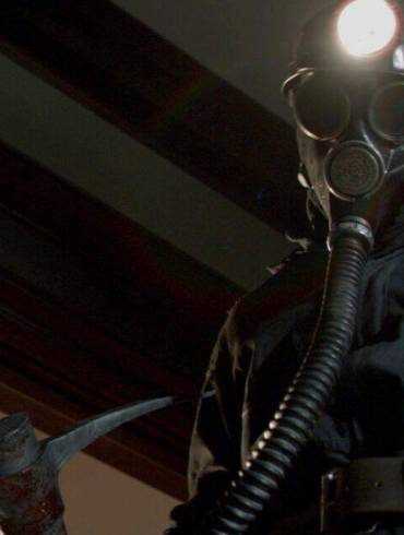 A terrifying figure in a dark uniform with a gas mask and axe in a dimly lit room.