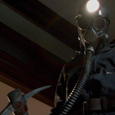 A terrifying figure in a dark uniform with a gas mask and axe in a dimly lit room.