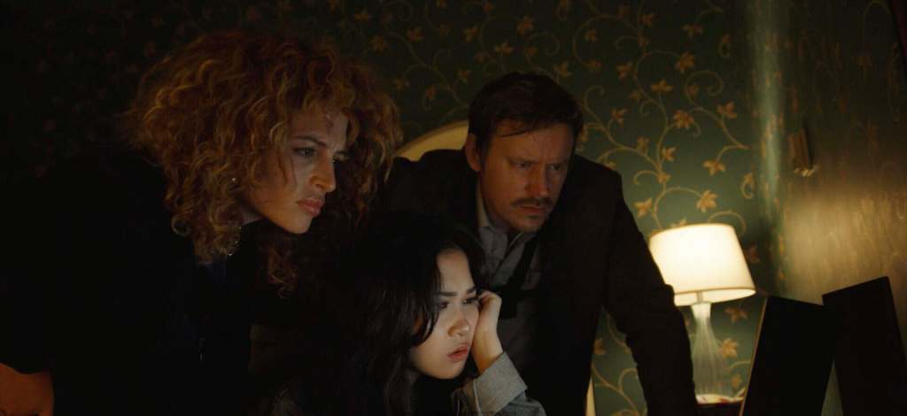 Three people look at a computer screen in a dimly lit room with green wallpaper.