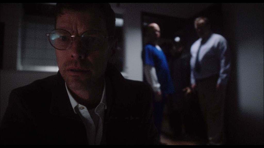 Dark scene with a man in glasses in foreground, three people in background in a dimly lit room.