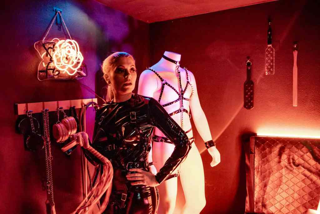 Leather-clad woman with whip in a BDSM-themed room with bondage gear and neon lighting.