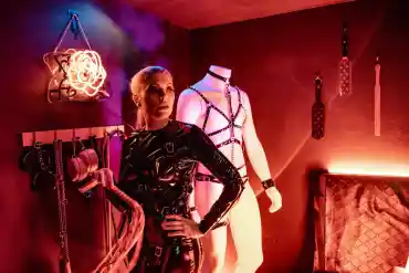 Leather-clad woman with whip in a BDSM-themed room with bondage gear and neon lighting.