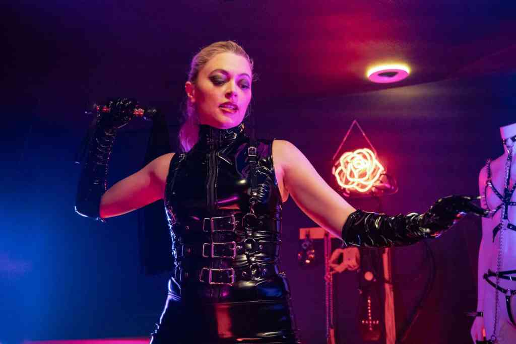 Woman in black latex outfit with gloves in a dark, neon-lit room.