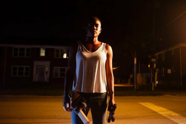 A woman standing alone at night on a deserted street with a flashlight and a book.