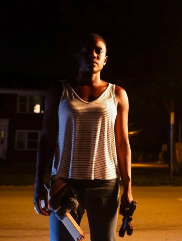 A woman standing alone at night on a deserted street with a flashlight and a book.