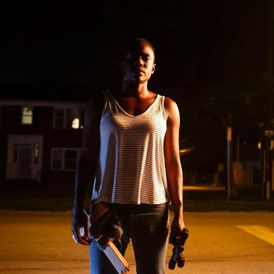 A woman standing alone at night on a deserted street with a flashlight and a book.