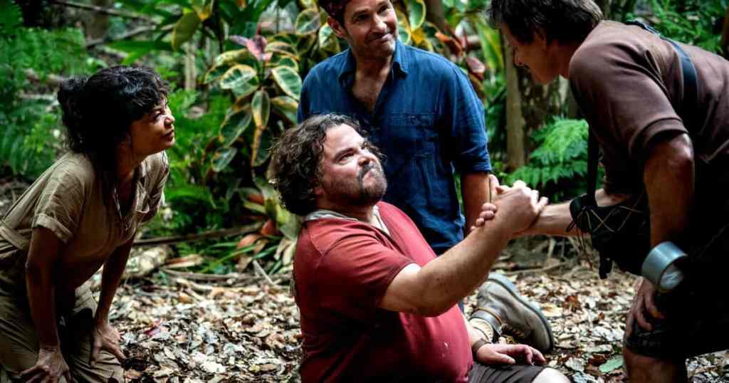 Group of four people in a jungle, one sitting and shaking hands with another.