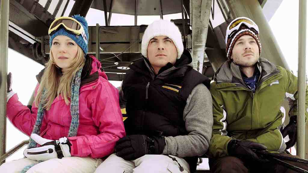 Three people sitting on a ski lift in winter gear, looking up.