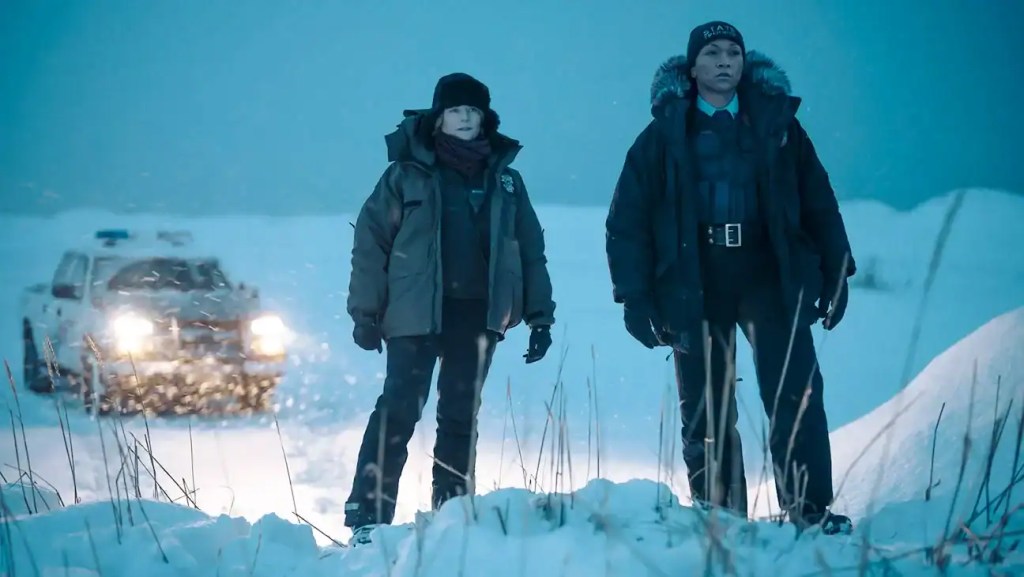 Two explorers in heavy winter gear stand in a snowy landscape with a vehicle in the background.