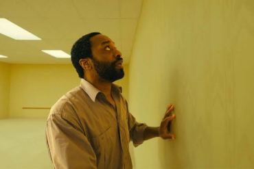Man in beige shirt touching yellow wall in empty room with fluorescent lights.