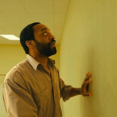Man in beige shirt touching yellow wall in empty room with fluorescent lights.