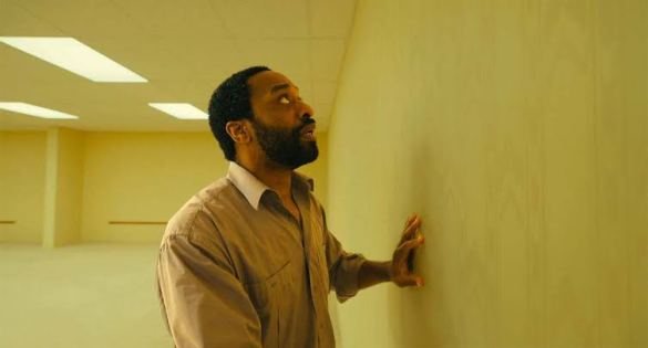 Man in beige shirt touching yellow wall in empty room with fluorescent lights.