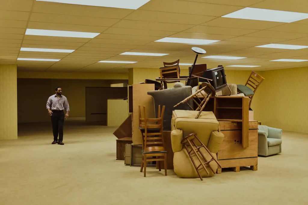 Cluttered room with furniture and a man in the background, eerie atmosphere.