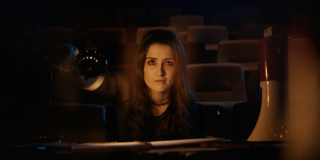 Young woman with long hair in a dark movie theater, looking at the camera.