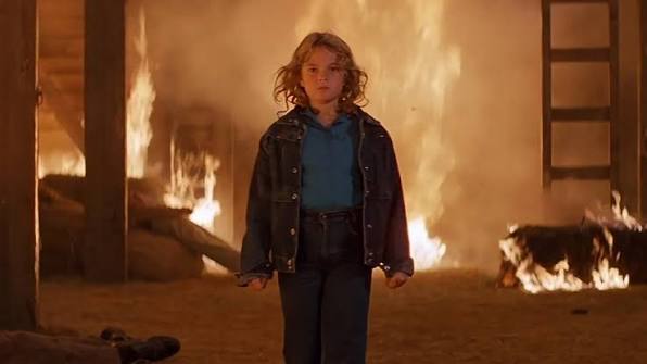 Young girl standing in a burning house with flames in the background.