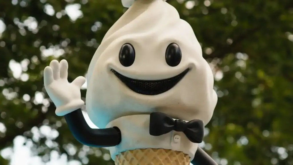Cute mascot character with a big smile, waving outdoors with green trees in background.
