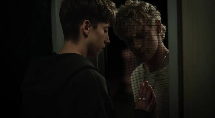 Two young men touching a glass window, looking at each other in a dark setting.