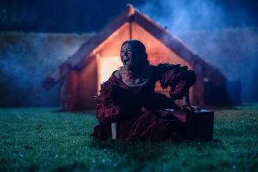 Creepy clown sitting on grass near a haunted house at night.