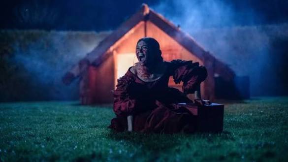Creepy clown sitting on grass near a haunted house at night.