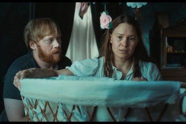 A woman and a man look into a baby cradle in a dimly lit room.