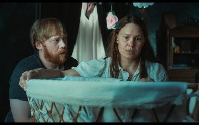 A woman and a man look into a baby cradle in a dimly lit room.