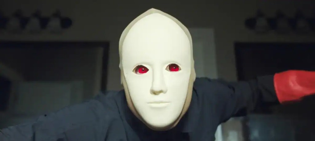 Person wearing a white mask with red eyes in a dark room.