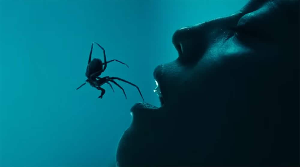 Close-up of a person with a spider near their open mouth in a dark setting.