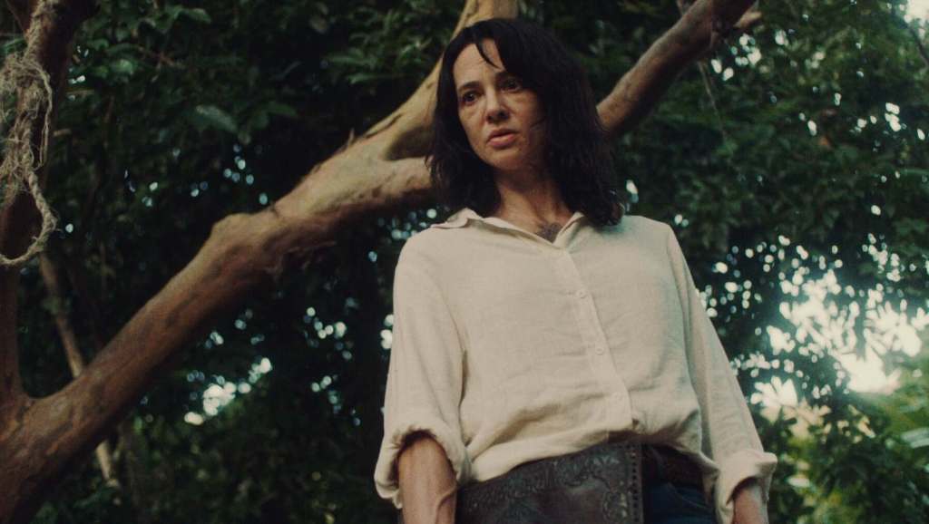Woman with dark hair in a white shirt outdoors near a tree.