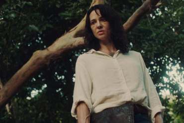 Woman with dark hair in a white shirt outdoors near a tree.