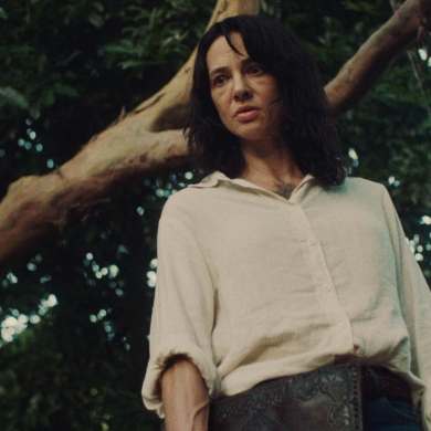 Woman with dark hair in a white shirt outdoors near a tree.