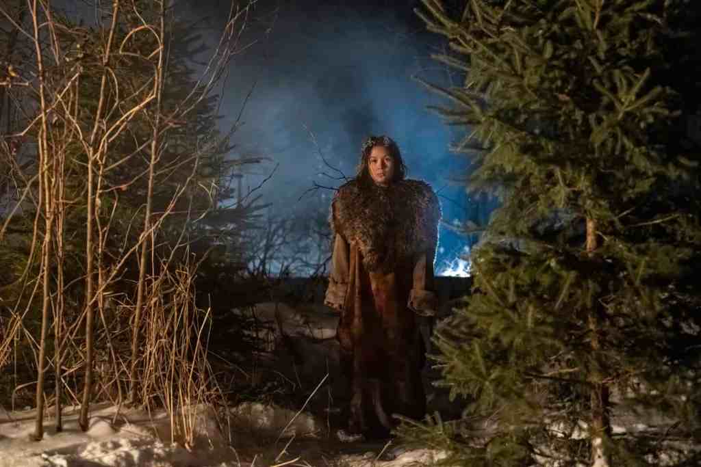 Woman standing in a snowy forest at night with fog and blue light in the background.