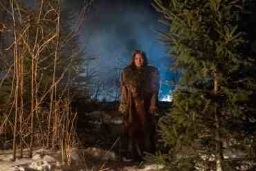 Woman standing in a snowy forest at night with fog and blue light in the background.