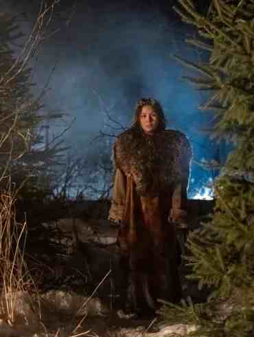 Woman standing in a snowy forest at night with fog and blue light in the background.