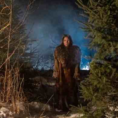 Woman standing in a snowy forest at night with fog and blue light in the background.
