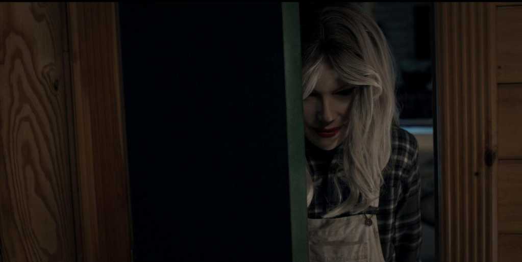Woman with blonde hair peeking from behind a door in a dimly lit room.