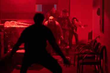 Group of people walking through a dimly lit, red-tinted room with chairs and tables.