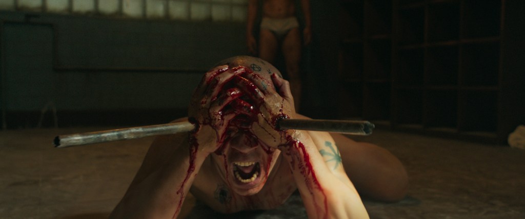 Horror scene of a man screaming, bloodied hands on head with barbell across forehead.