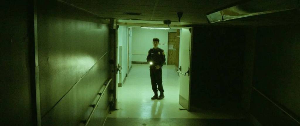 Eerie hospital corridor with a lone figure standing at the end in dim lighting.