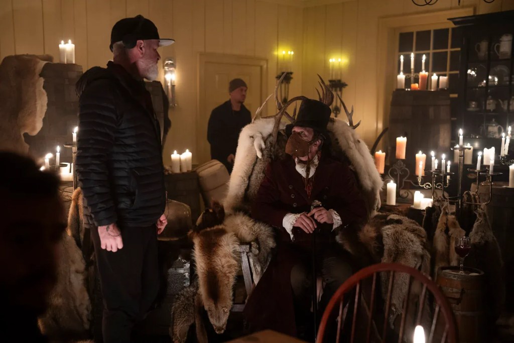 Actors in a dimly lit room with candles, one in a throne-like chair wearing a fur cloak.